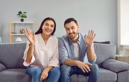 Happy smiling couple waving hands looking at camera. Cheerful husband and wife making online video call. Couple sitting on couch at home enjoying virtual meeting, video call chat. Online communicationの写真素材