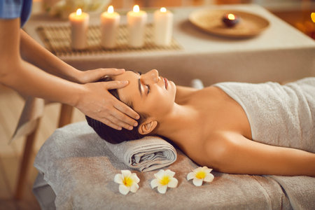 Close up portrait of a beautiful young woman lying with closed eyes and having face or head massage in spa. Woman relax enjoying health therapy treatment. Wellness and beauty day concept.の写真素材