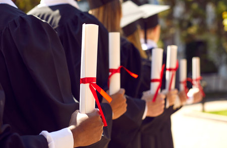 College or university graduates at outdoor graduation ceremony. Diverse group of students standing in row and holding diploma scrolls tied with red ribbons. Concept background. Cropped closeup shotの写真素材