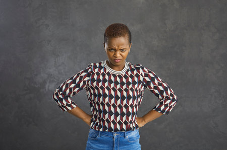 Frustrated, angry, and gloomy African-American woman is unhappy and needs an explanation. Woman holds her hands on her sides and angrily looks at the camera while standing on a gray background.の写真素材
