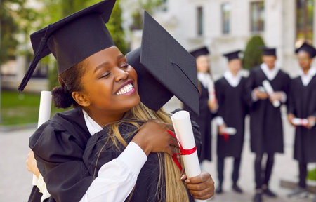 Congrats, grad. Proud of you. Happy, joyful university graduates celebrate graduation together. Beautiful Afro American student girl in square black cap holding scroll, hugging her friend and smilingの写真素材