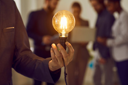 Creative thinking. Glowing electric light bulb in hand of unknown smart and successful businessman dressed in suit. Close up of Edison light bulb on background of people. Concept of new idea.の写真素材