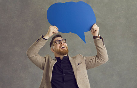 Excited young man in suit and hipster glasses showing cardboard thought bubble isolated on grey...