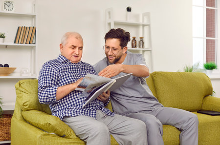 Old man and an interested retirement home worker looking through a photo album together. Two men sitting on the sofa and looking at pictures in a photobook or family photo album. Senior care conceptの写真素材