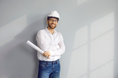 Happy young man architect or construction engineer in white shirt, safety hard hat, and eyeglasses standing by light grey house wall, holding paper blueprint plan roll, looking at camera, and smilingの写真素材