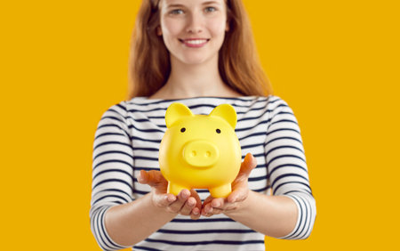 Cropped shot of a happy beautiful young Caucasian woman standing on a yellow studio background, holding a yellow piggy bank and smiling. Concept of saving moneyの写真素材