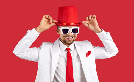 Portrait of smiling young man in suit, hat and glasses isolated on red studio background. Happy millennial male showman or performer make tricks and jokes for audience. Entertainment.の写真素材