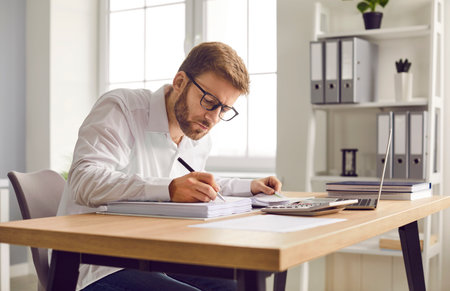 Busy financial accountant working at his office desk. Young man in a white shirt and glasses sitting at his office table, doing business paperwork, and using a modern laptopの写真素材