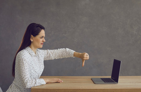 Side profile view lady with discontent face expression sitting at desk, giving thumbs down at laptop...