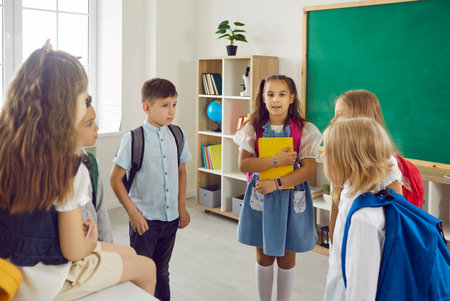 Schoolchildren talking together in class after lessons holding folders and bags. Boys and girls elementary school students standing and sitting on desk in round. Education, communication of children.の写真素材