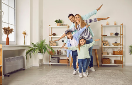 Happy smiling family having fun with their two kids boy and girl in the living room at home. Couple with cheerful children in pilots glasses raising arms to the sides like flying and playing plane.の写真素材