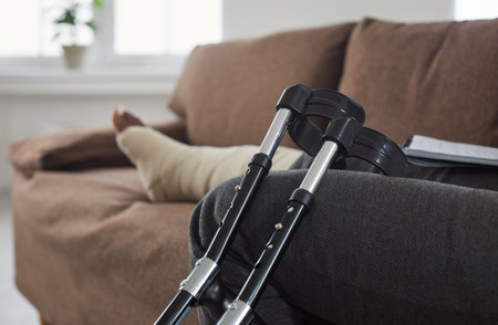 Close-up of picture of crutches against background of sitting african american woman resting at home sitting on the sofa in the living room. Rehabilitation after leg injury, severe bruise or sprain.の写真素材