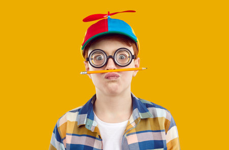 Studio portrait of funny preteen boy who is fooling around holding pencil as mustache. Child boy in funny glasses and cap holding balancing pencil between lips and nose isolated on orange background.の写真素材