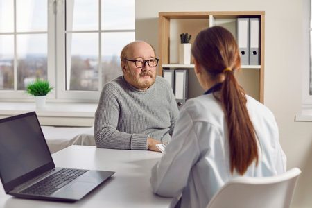 Senior patient comes to the clinic for a professional medical consultation. Bald elderly man in glasses sitting at a desk with a doctor and listening to young medical specialists adviceの写真素材