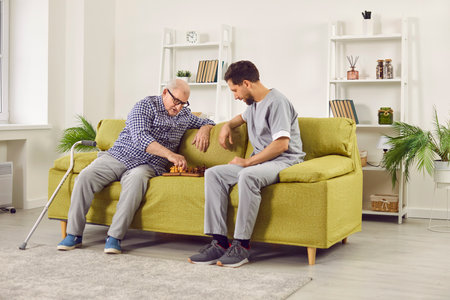 Senior man playing chess with nurse in retirement home. Old man in glasses sitting on sofa together with young man in uniform, playing chess, and thinking which move to make. Senior care conceptの写真素材