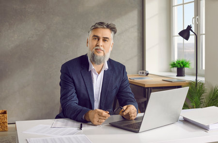 Portrait of a serious handsome bearded mature business man in suit holding glasses working at the desk on his workplace with a laptop at office or at home and looking confident at the camera.の写真素材