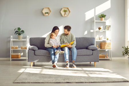 Smiling , happy mother and father with charming child engaged in educational activities. Loving family with their young child read a book together, sitting at home on cozy sofa.の写真素材