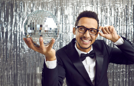 Positive young african american man in suit holding disco ball preparing for party or celebration. Happy man looks at silver disco ball in his hand standing on background of foil silver curtain.の写真素材