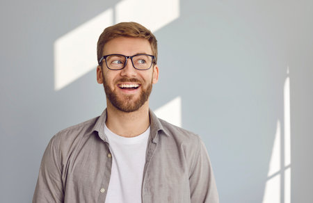 Portrait of smiling young bearded handsome man looking to the side with his eyes and open mouth wearing white t-shirt and beige shirt isolated on grey wall background with copy space.の写真素材