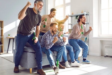 Male football fans watching sport match on TV together at home. Group of emotional friends sitting on sofa with beer bottles and celebrating victory of their favorite team. Friendship, entertainmentの写真素材