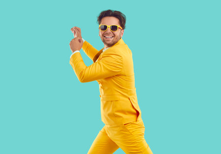 Cheerful stylish man having fun dancing to moving and rhythmic music on light blue background in studio. Cool Caucasian smiling man in yellow suit and sunglasses dancing energetically. Isolated.の写真素材
