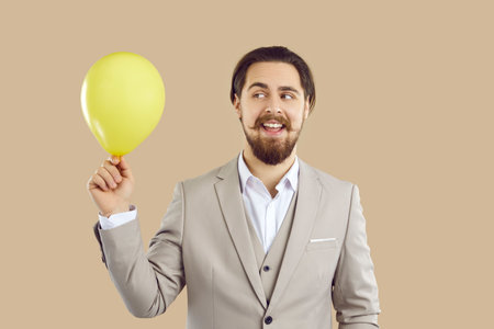 Handsome stylish young man in three-piece suit holding yellow inflatable balloon on beige background. Cheerful fashionable man with beard and mustache who looks funny at balloon in his hand.の写真素材