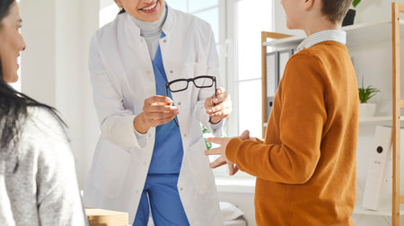 Doctor gives new glasses to child patient. Friendly smiling woman optometrist or ophthalmologist gives eyeglasses to little boy who came to see her with his mother. Ophthalmology and optometry conceptの写真素材