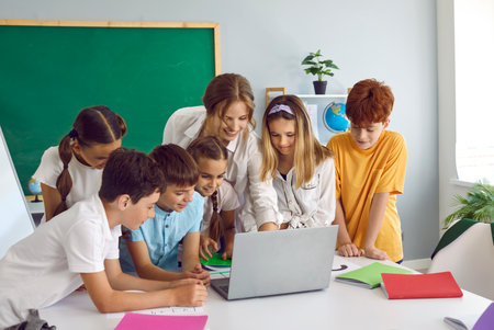 Smart children pupil boys and girls stand in front of laptop with woman teacher and view media material or online presentation during lesson in primary school classroom. Curiosity, educationの写真素材
