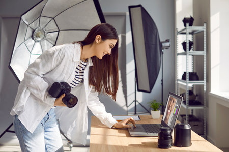 Portrait of a young smiling girl photographer at her workplace with camera and lenses retouching female portrait photo on a laptop in production studio with professional light equipment.の写真素材