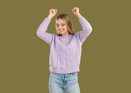 Happy beautiful funny blonde young girl dressed in a warm sweater and light blue jeans is fooling around and dancing with her hands up. Studio photo of beautiful blonde on isolated background.の写真素材
