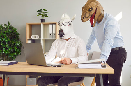 Two funny business men wearing animal masks standing at their workplace with laptop and having discussion in office, considering new projects or startups, analyzing company. Fools day concept.の写真素材