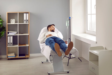 Young man attached to intravenous drip lying on bed in medical clinic. Male patient getting vitamin IV infusion drip therapy in hospital room. Medical treatment, maintenance of immune systemの写真素材