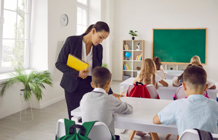 Female teacher holding yellow folder helps caucasian schoolkids to finish they lesson indoors. Interior of light modern school classroom, view from back desks. European education system.の写真素材