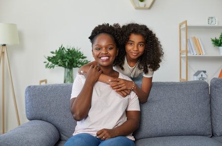 Portrait of happy family on sofa at home. Indoor shot of cheerful mother and child. Beautiful young African American mom and daughter hugging, looking at camera and smilingの写真素材