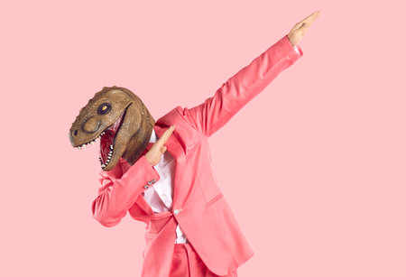Cheerful young man in dinosaur rubber mask makes dab dance gesture isolated on pink background. Man with lizard mask on his head dressed in formal pink suit is having fun and shows dabbing movement.の写真素材