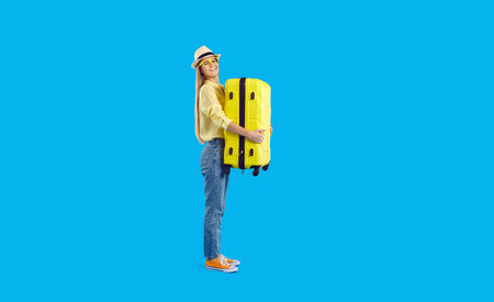 Funny cheerful girl traveler with yellow travel suitcase isolated on light blue background. Stylish young woman holding suitcase and smiling at camera. Summer holiday concept. Banner. Full length.の写真素材