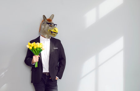 Adult man wearing fashionable suit and funny bizarre eccentric silly freak absurd surreal masquerade animal donkey mask with glasses holding bunch of flowers on blank advertising space wall backgroundの写真素材