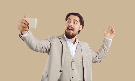 People lifestyle. Cheerful stylish man in suit takes selfie, records video or talks online on beige background. Smiling active millennial Caucasian man in three-piece suit uses modern smartphone.の写真素材