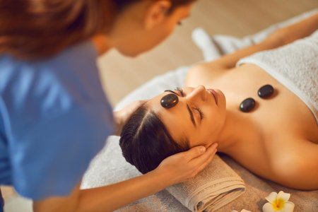 Beautician making face massage to female client in spa salon. Beautiful girl lying on massage bed on with closed eyes enjoying spa procedure and relaxing. Facial beauty treatment, body care, wellnessの写真素材