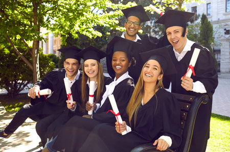Group of laughing multicultural people in graduation gowns and caps with diplomas graduate...