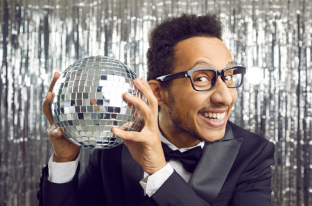 Portrait of smiling young African American man on glitter shiny background hold disco ball. Happy millennial black guy in glasses and formal suit wear with discoball celebrate party in night club.の写真素材