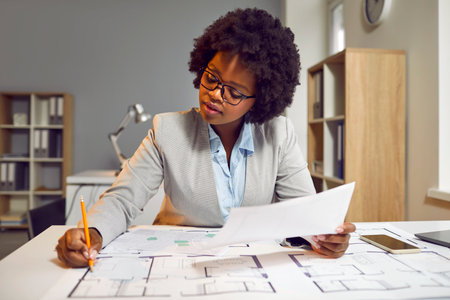Female architect developing and drawing architecture project on paper. Young Afro American woman in glasses sitting at office desk, holding pencil, and working with modern house blueprint floor plansの写真素材