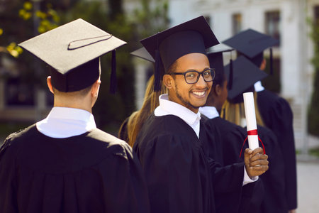 Half turned smiling indian graduate man in glasses in graduation clothes looking at camera and smiling on ceremony. Another graduates people going back to camera. Graduation from college, university.の写真素材
