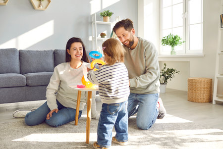 Young mother, father and their little daughter spend time at home in cozy childrens room. Parents are sitting on floor and playing with different toys with their child. Family holiday concept.の写真素材
