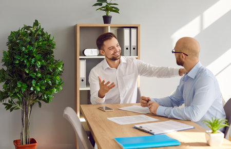 Smiling businessman touching shoulder of partner in office. Two businessmen, partners sitting at desk negotiating business strategy, sharing ideas, discussing new project, having informal conversationの写真素材