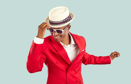 Time for party. Happy refined and stylish african man having fun dancing on pastel light blue background. Joyful positive man in red suit, white shirt, sunglasses holding on to hat having fun at partyの写真素材