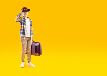 Happy teenage boy with suitcase looking through binoculars. Cheerful casually dressed boy boy holding modern suitcase over isolated copy space studio background. Happy child dreaming about adventuresの写真素材