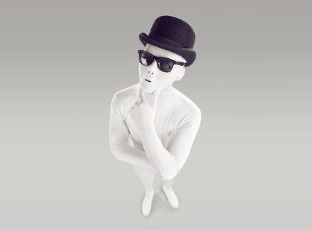 Hmm, well, let me think. Unusual incognito adult young man wearing white bodysuit costume, sunglasses and black color bowler hat trying to answer question. High angle view from above, gray backgroundの写真素材
