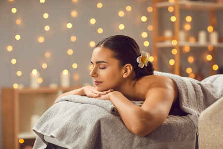 Beautiful woman lying on massage bed with closed eyes. Relaxed happy brunette girl enjoying spa treatment procedure in spa salon. Beauty, spa treatment, skin care, wellbeingの写真素材