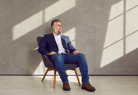 Portrait of successful businessman in smart casual suit. Relaxed confident mature business man in classic blue jacket, shirt, jeans, and brown shoes sitting on chair on grey office wall backgroundの写真素材